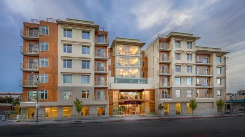Modern six-story apartment building with large windows, balconies, and a well-lit glass entrance, situated along a street with young trees and a cloudy sky in the background.