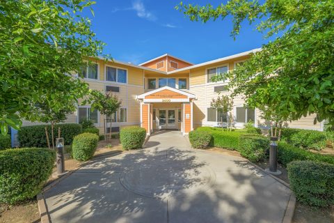 A two-story beige and yellow building with white trim, surrounded by green trees and shrubs, and a curved concrete walkway leading to the main entrance under a small awning labeled 2400.