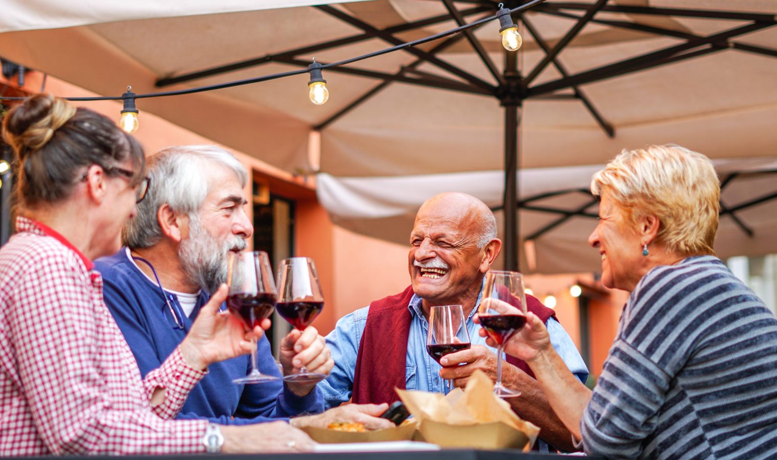 Four older adults sit around an outdoor table under string lights, smiling and clinking glasses of red wine, enjoying each other’s company on a sunny day.