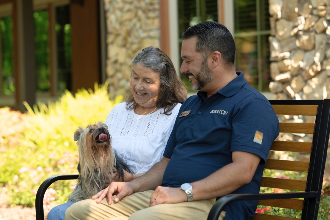 An older woman and a man sit smiling on a bench outdoors with a small, long-haired dog. The woman wears a white blouse, and the man wears a navy shirt. The dog sits happily on the woman’s lap.