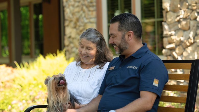 An older woman and a man sit smiling on a bench outdoors with a small, long-haired dog. The woman wears a white blouse, and the man wears a navy shirt. The dog sits happily on the woman’s lap.