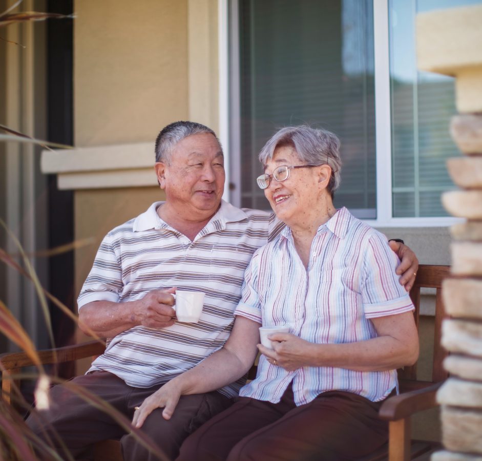 An elderly couple sits together on a bench outside, smiling and holding white mugs. The man has his arm around the woman. They appear relaxed and happy, enjoying each others company near a windowed house.