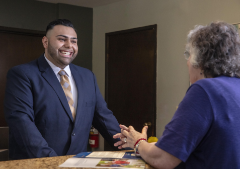 A man in a suit smiles and talks with an older woman across a counter. The woman is wearing glasses and a blue shirt, and there are brochures on the counter between them.