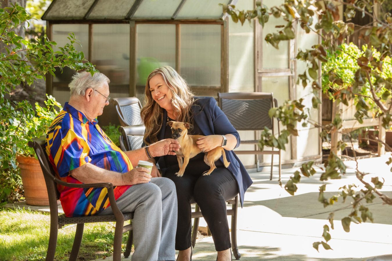 An older man and a woman sit on a bench outdoors, smiling and petting a small dog. The man holds a coffee cup, and they are surrounded by greenery near a greenhouse.