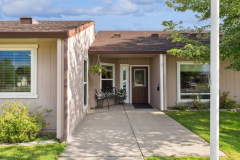 Single-story beige house with a brown roof, white-trimmed windows, and a central entrance. There’s a bench and hanging flower basket near the front door, and green lawn with small bushes on either side of the walkway.