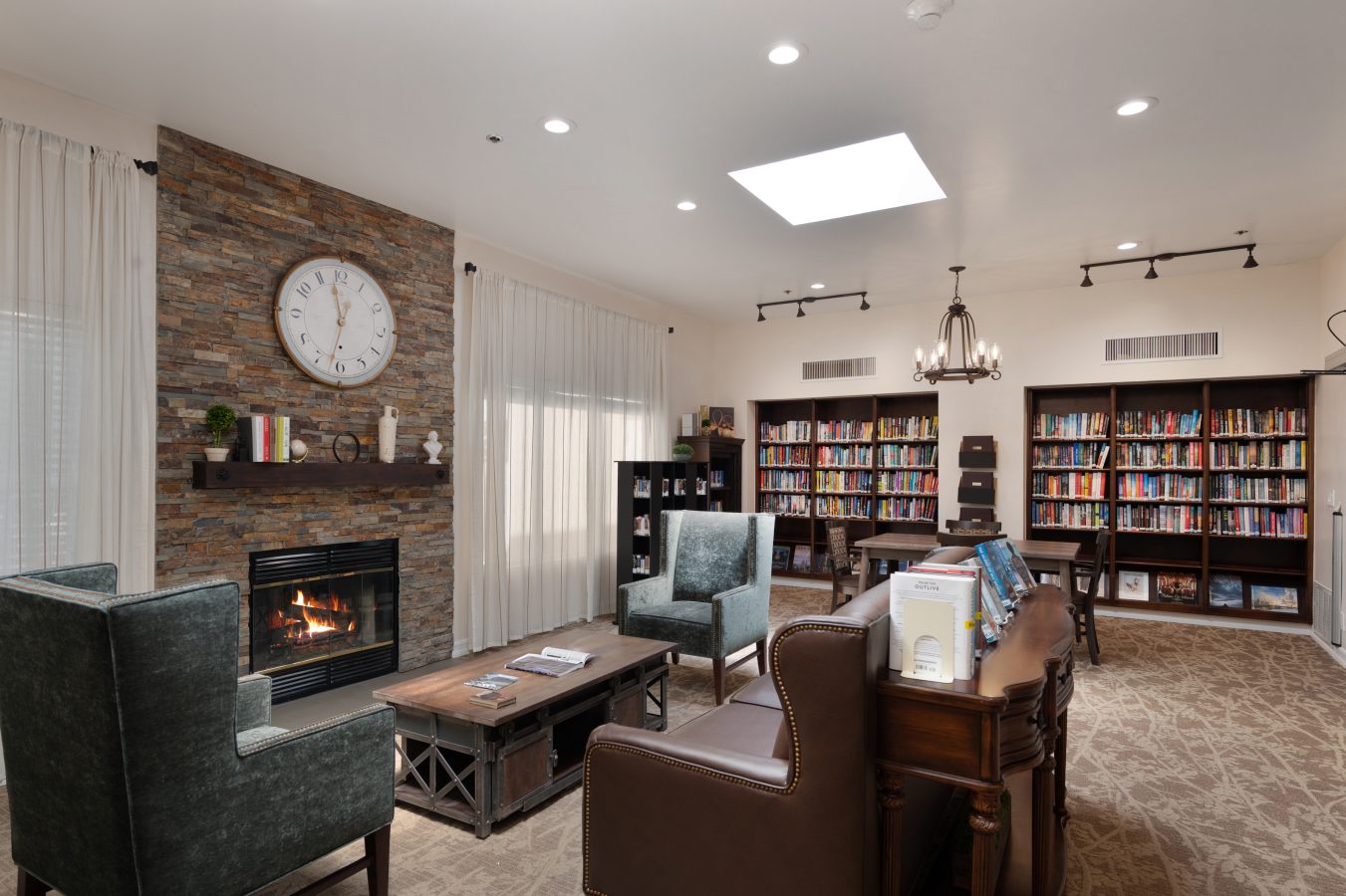 A cozy library with bookshelves, a stone fireplace, armchairs, a wooden coffee table, and a large clock above the mantel. Warm lighting and natural light fill the room, creating an inviting reading space.
