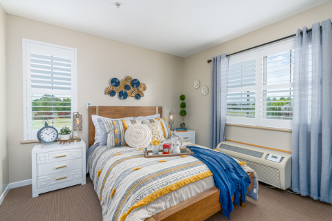 A cozy bedroom with a wooden bed, colorful patterned bedding, decorative pillows, and a breakfast tray. Two nightstands hold lamps and decor. Large windows with white shutters and blue curtains let in natural light.