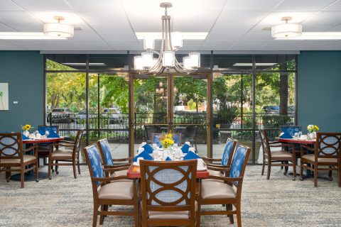 A bright dining room with four tables set for meals, decorated with sunflowers. Large windows reveal a green, leafy outdoor view. Modern light fixtures hang from the ceiling, and wooden chairs surround each table.