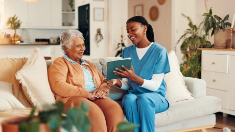 A nurse in blue scrubs sits on a couch with an older woman, smiling and holding a book together in a cozy, well-lit living room.