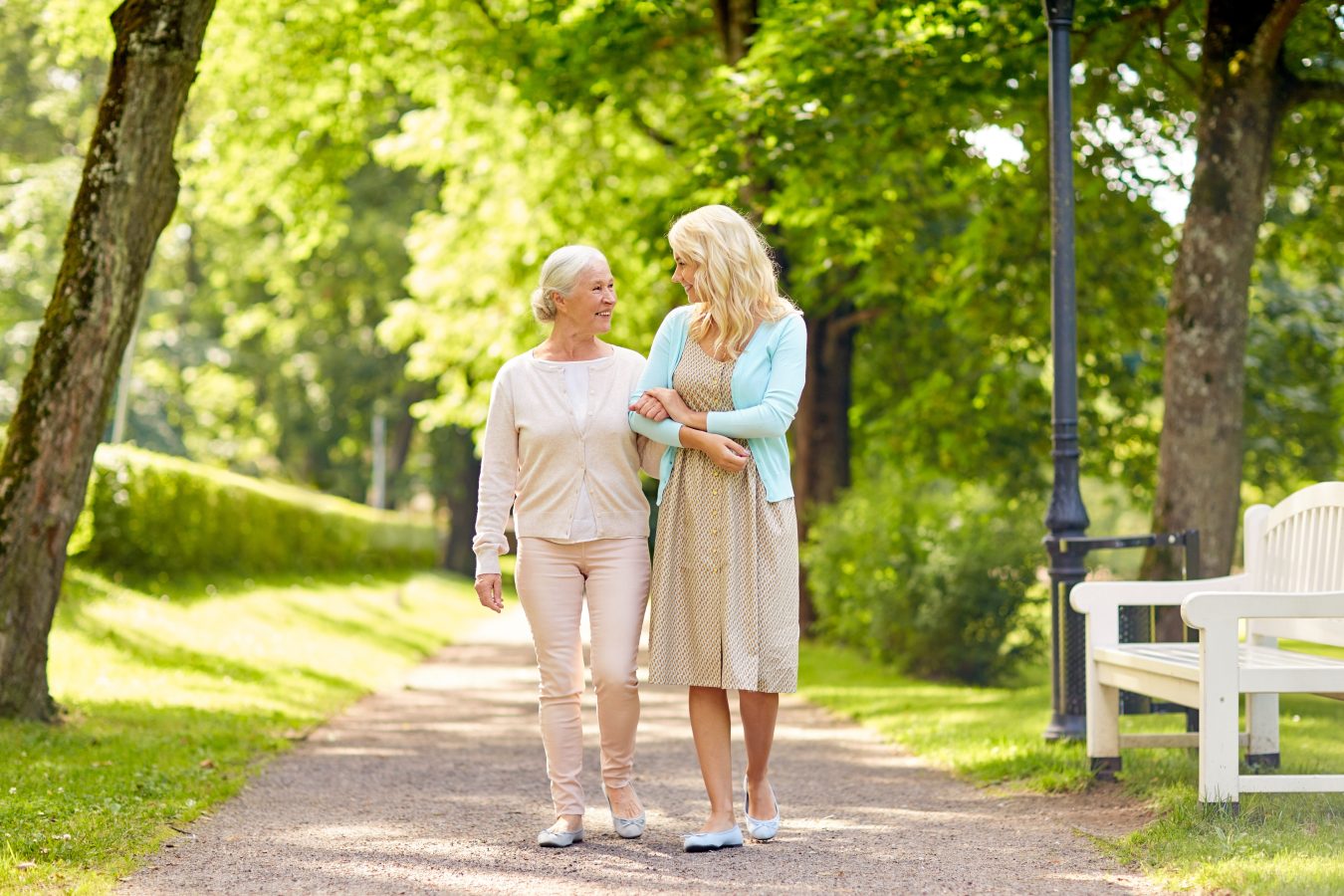 Two women, one older and one younger, walk arm-in-arm along a tree-lined path in a park on a sunny day, smiling at each other. A white bench is visible on the right.