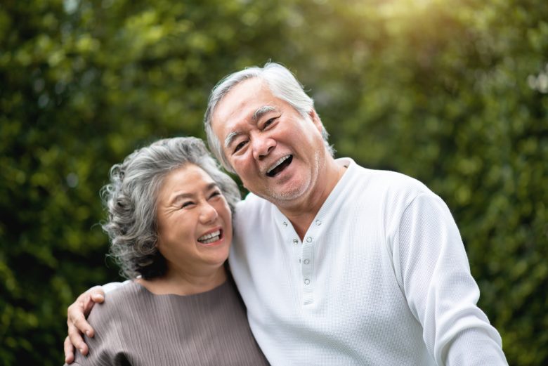 An elderly couple smiles and embraces outdoors, standing in front of green foliage. Both appear happy and joyful, with the man wearing a white shirt and the woman in a brown top.