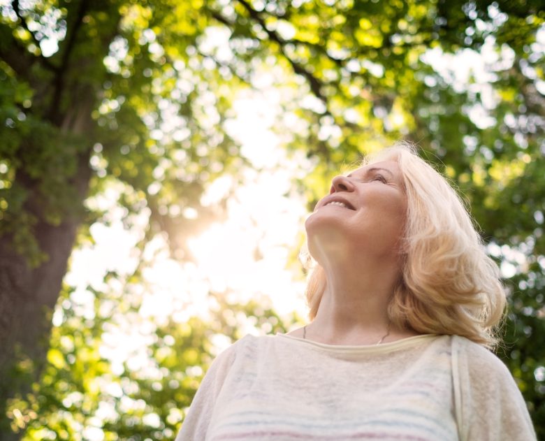 A smiling woman with blonde hair looks up, standing outdoors under bright sunlight and green, leafy trees. The sun shines through the branches, creating a warm and peaceful atmosphere.