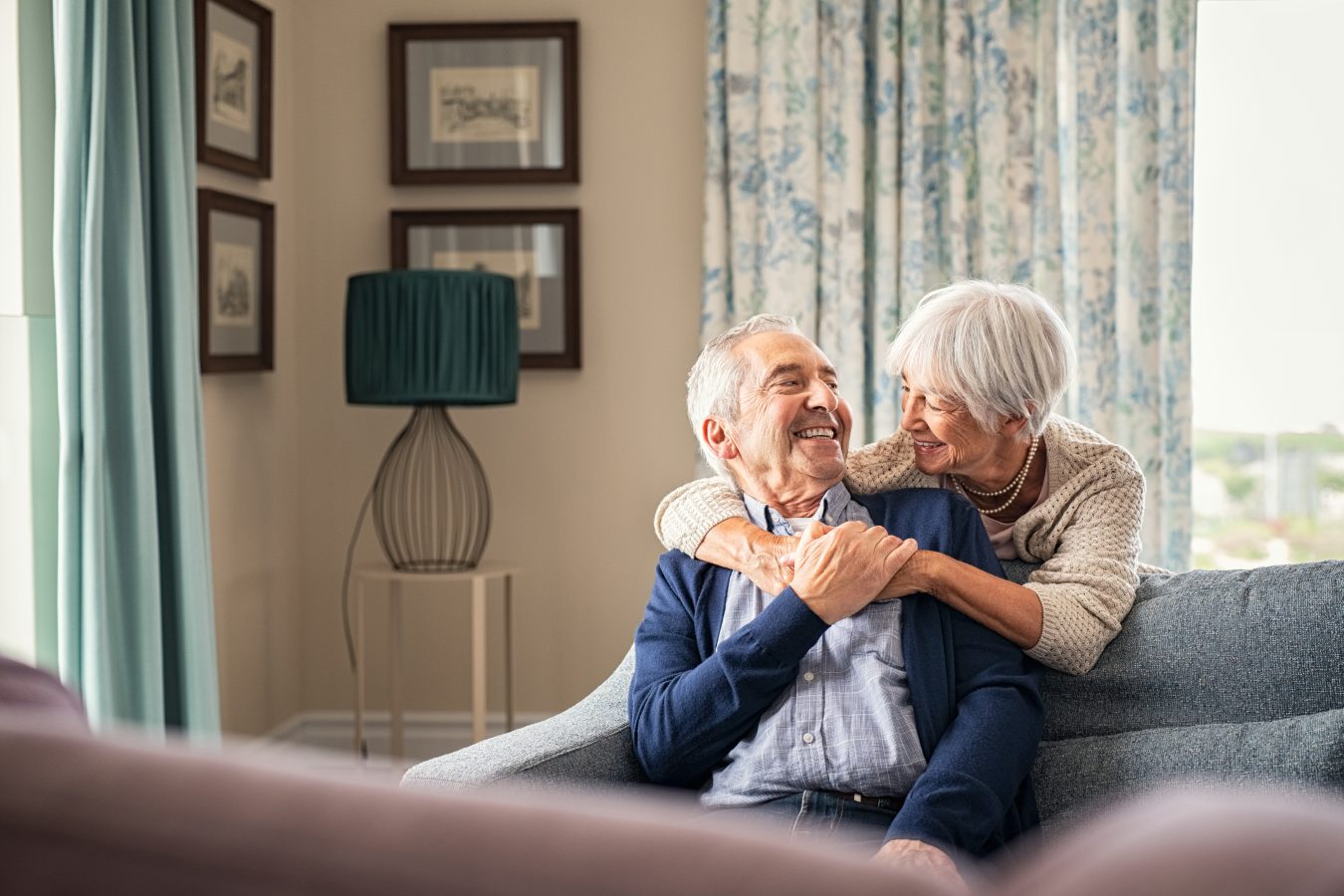 An elderly couple sits on a sofa at home, smiling and embracing each other warmly. The room is softly lit with patterned curtains, framed pictures on the wall, and a lamp in the background.