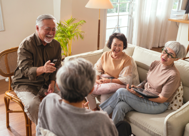 Four older adults sit together in a bright living room, smiling and laughing as they chat. One person gestures while others listen, creating a warm and friendly atmosphere.