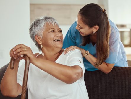 An older woman with short gray hair sits holding a cane and smiles at a young caregiver who stands behind her, gently touching her shoulders. Both women look happy and are sharing a warm moment in a bright room.
