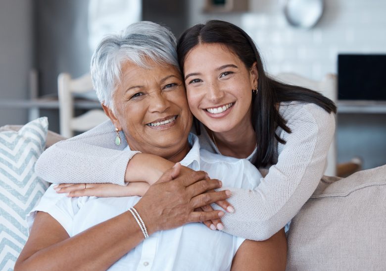 A young woman with long brown hair hugs an older woman with short gray hair from behind. Both are smiling warmly while sitting on a couch in a bright, cozy living room.
