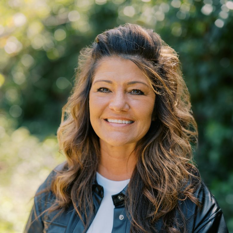 A woman with wavy brown hair and highlights smiles outdoors. She is wearing a black jacket over a white top, with a green, leafy background blurred behind her in natural light.