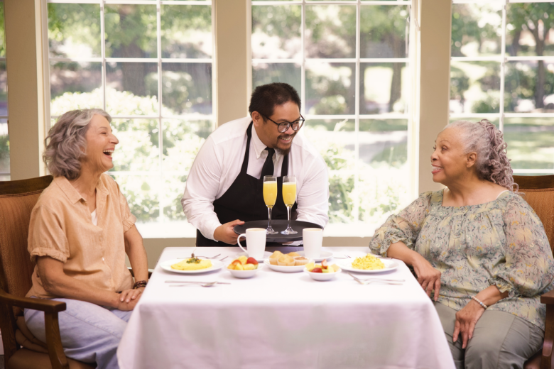 Two older women sit at a table with breakfast foods and drinks, smiling and talking as a waiter stands between them, also smiling. Large windows behind them show a green outdoor view.