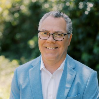 A middle-aged man with short, curly gray hair and glasses smiles while wearing a light blue blazer and white shirt, standing outdoors with greenery blurred in the background.