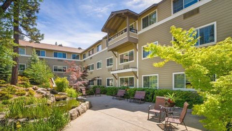 A modern, three-story beige building with large windows overlooks a landscaped courtyard featuring patio seating, lush greenery, small trees, and a pond with rocks under a blue sky.