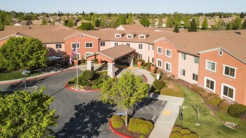 Aerial view of a large, two-story tan building with a red-tiled roof, landscaped grounds, American flag, circular driveway, and surrounding trees in a suburban neighborhood.