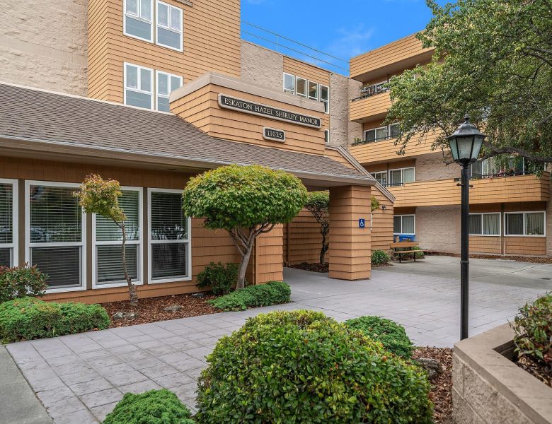 Entrance to a tan, multi-story building with large windows, a sign reading “Eskaton Hazel Shirley Manor,” trimmed bushes, trees, and a lamp post along a paved walkway under a blue sky.