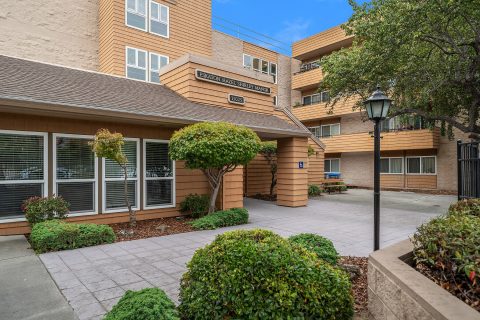 A three-story tan apartment building with large windows and balconies, landscaped with shrubs and trees, featuring a covered entrance with a sign reading EASTON TOWER SENIOR HOUSING above the doorway.