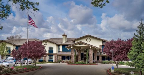 A large building with an American flag out front, surrounded by landscaping with pink-blossomed trees, under a partly cloudy sky. Several cars are parked along the circular driveway.