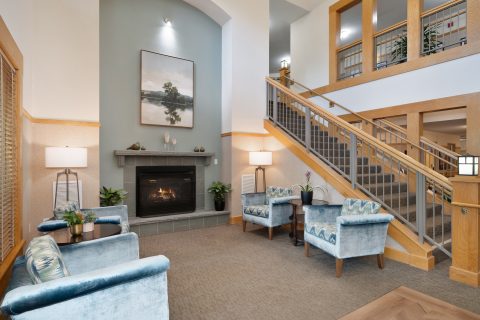 A cozy lobby with two blue armchairs, a fireplace, and a painting above the mantel. A wooden staircase with metal railings leads upstairs. The space is warmly lit with table lamps and natural light.