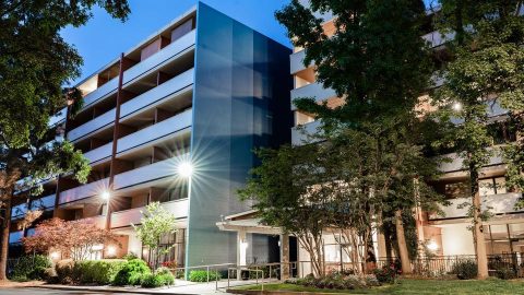 A modern multi-story apartment building with balconies, illuminated by outdoor lights at dusk, surrounded by trees and landscaped greenery.