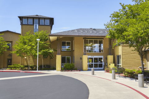 A modern building with large windows and tan siding, surrounded by green trees and landscaping, under a clear blue sky. A curved driveway and sidewalk lead to the main entrance.
