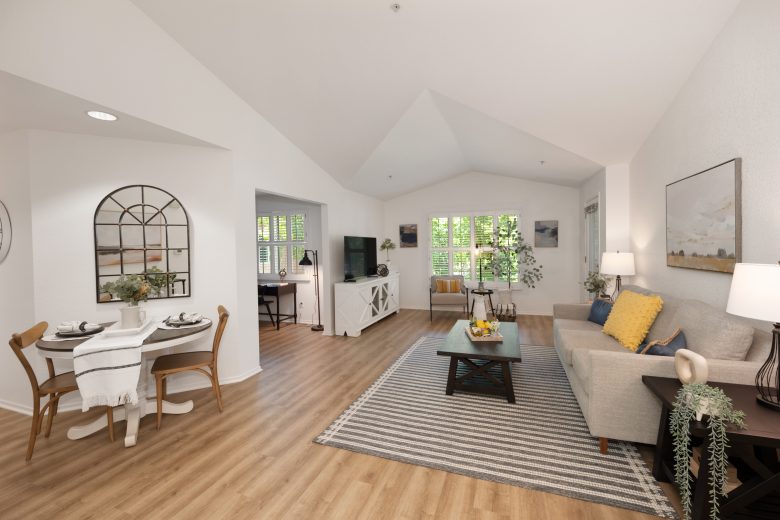 A bright, open living and dining area with light wood floors, white walls, a small dining table, a gray sofa with yellow pillows, a striped rug, and large windows letting in natural light.