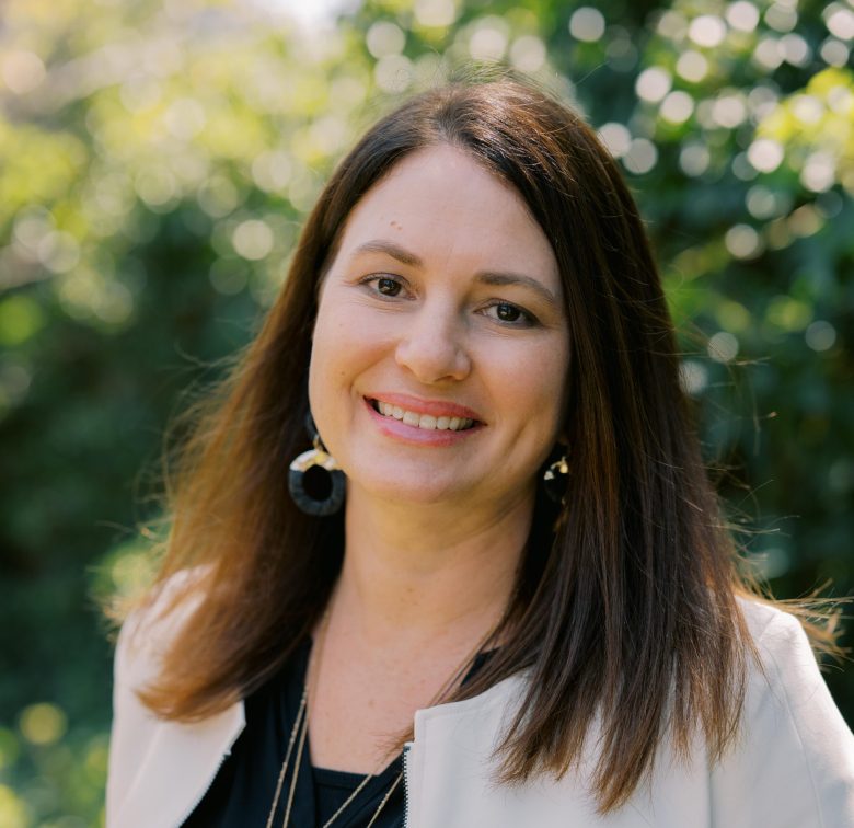 A woman with straight brown hair, wearing a light-colored blazer, black top, and large round earrings, smiles outdoors with greenery and sunlight softly blurred in the background.