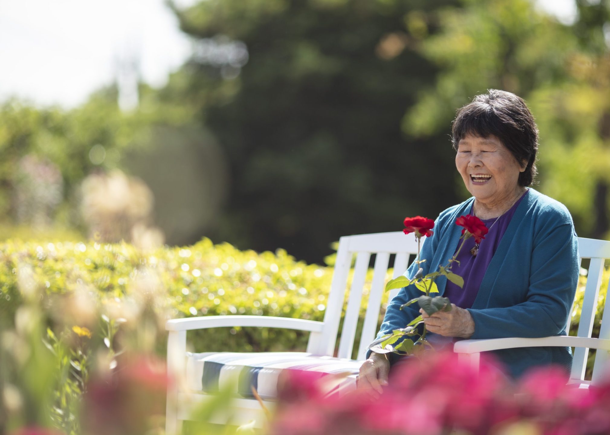 An elderly woman sits on a white bench in a sunny garden, smiling and holding a red rose, with greenery and flowers in the background.