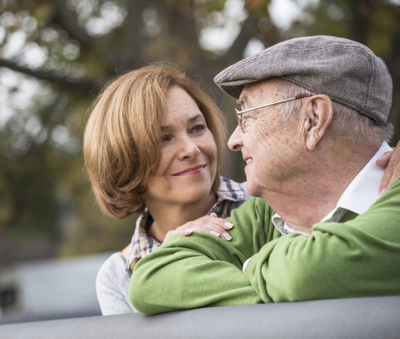 A smiling older man wearing a flat cap and green sweater sits closely with a woman who has short hair and a plaid shirt. They look at each other affectionately outdoors.