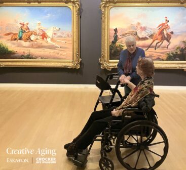 Two older women, one in a wheelchair and one with a walker, enjoy viewing large, colorful paintings in a bright museum gallery—an inspiring scene of Creative Aging with Crocker Art surrounded by ornate gold-framed artwork.