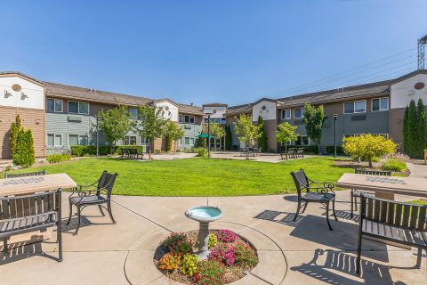 A sunny courtyard with green grass, blooming flowers, benches, tables, and a small birdbath, surrounded by two-story residential buildings with beige and green exteriors.
