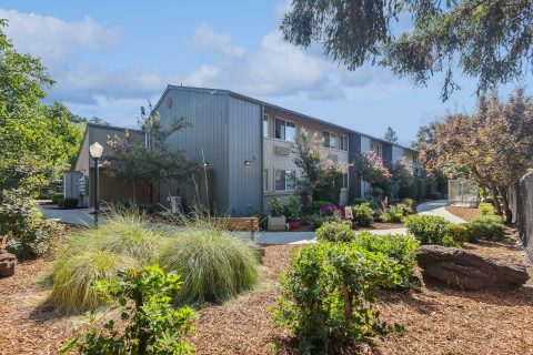 A two-story gray apartment building surrounded by landscaped gardens, shrubs, and trees, with a pathway leading to the entrance under a partly cloudy sky.