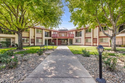A wide concrete pathway leads to the entrance of a two-story, red and cream apartment building, surrounded by green lawns, trees, benches, and landscaped gardens with gravel and flowers.