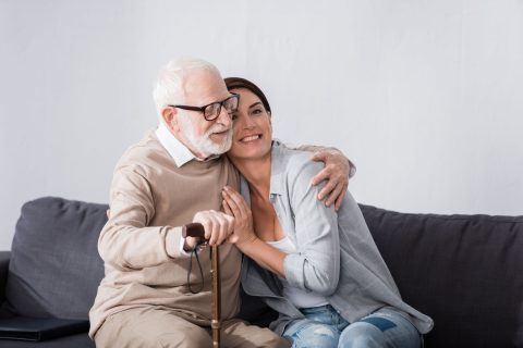 An elderly man with white hair and glasses, holding a cane, sits on a couch and hugs a smiling woman who leans into him affectionately. Both look happy and content.