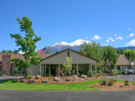 A single-story house with an American flag, surrounded by green grass, trees, and landscaping. Snow-capped mountains and a clear blue sky are visible in the background.