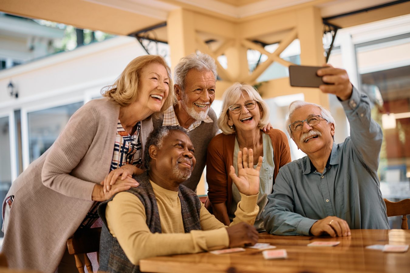 A group of five smiling older adults gather around a table, posing for a selfie. One person holds a smartphone, while another waves at the camera. They appear happy and relaxed in a bright, indoor setting.