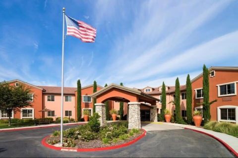 A large building with terracotta-colored walls and tall windows, an arched entrance, potted plants, and a U.S. flag waving on a pole in the foreground under a blue sky with wispy clouds.