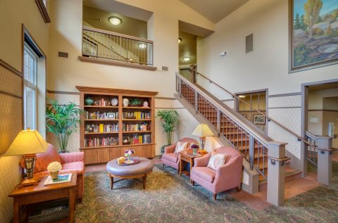 A cozy living area with two pink armchairs, a round ottoman, a wooden bookshelf filled with books, plants, and lamps. There’s a carpeted staircase leading to an upper level and a large window letting in natural light.