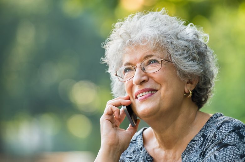 Smiling older woman with curly gray hair and glasses talks on a mobile phone outdoors, with sunlight and greenery in the blurred background.