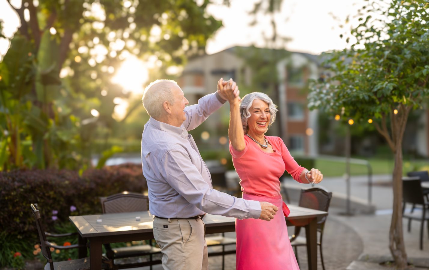An elderly couple dances joyfully outdoors at sunset, smiling and holding hands. The woman wears a pink dress, and the man wears a light shirt and khakis. They are surrounded by greenery and outdoor furniture.