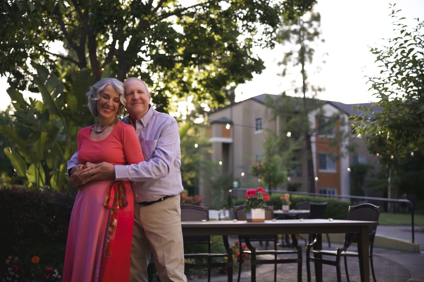 An older couple stands smiling and embracing in a lush garden, with an outdoor dining table and apartment buildings in the background. The woman wears a pink dress and the man wears a light shirt and khakis.