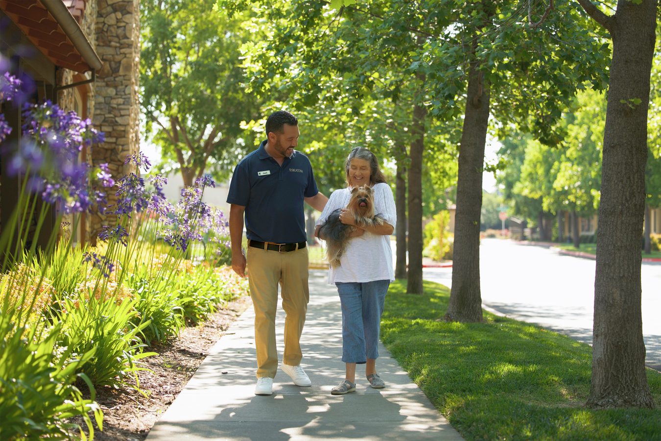 A man and an older woman walk together on a sunny, tree-lined sidewalk. The woman holds a small dog, and they are both smiling, surrounded by green plants and purple flowers.