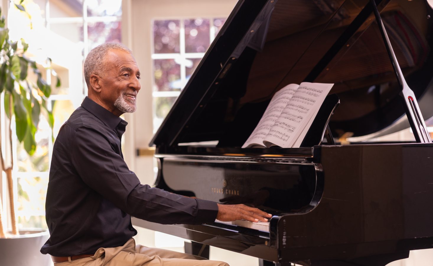 An older man smiles while playing a grand piano with sheet music open, seated in a bright room with large windows and a potted plant in the background.