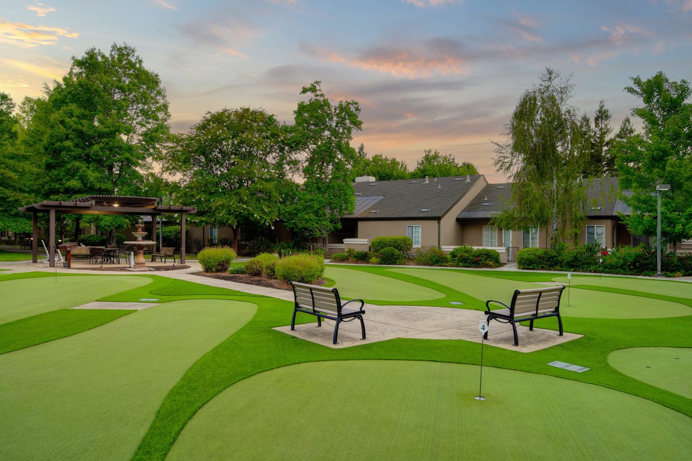 Two benches overlook a well-maintained putting green, surrounded by trees and a landscaped garden, with a pergola seating area and residential buildings in the background at sunset.